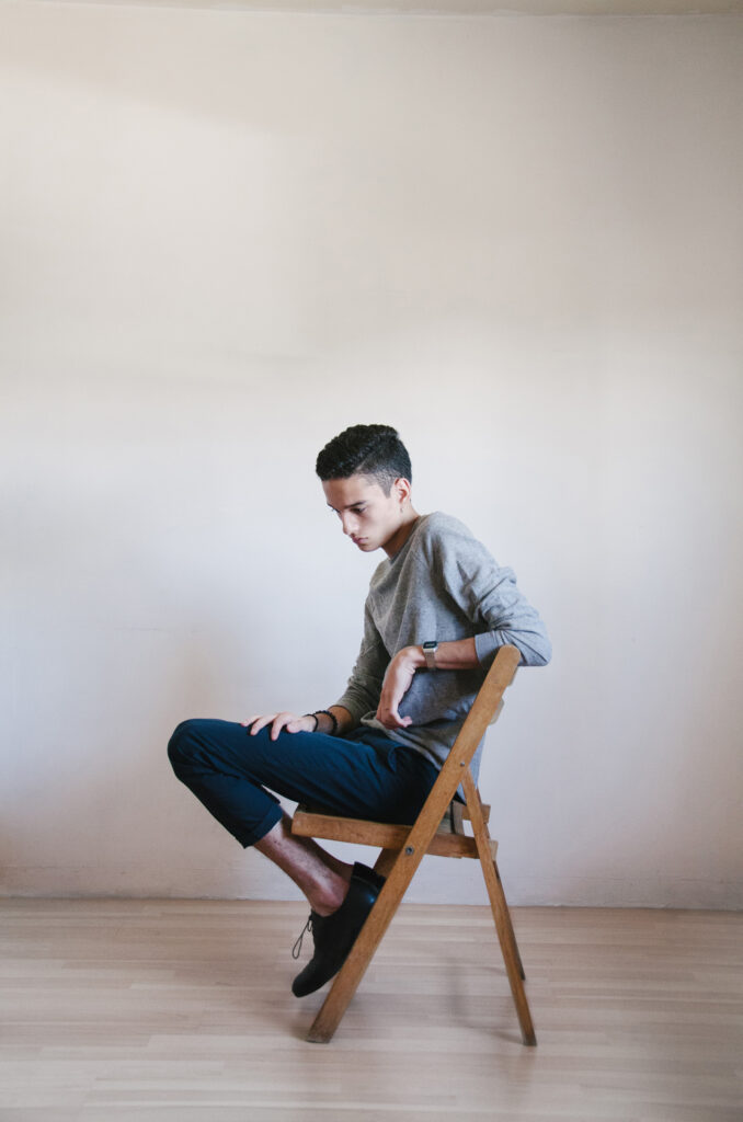 Victor Zepeda sits contemplatively on a wooden chair against a plain white wall wearing a gray sweater blue trousers and black shoes.